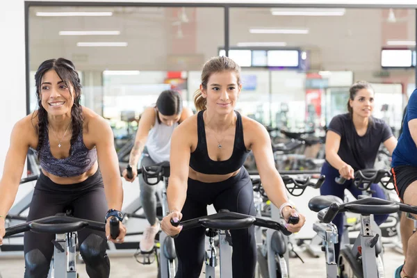 Smiling female clients riding exercise bikes while looking away in health club