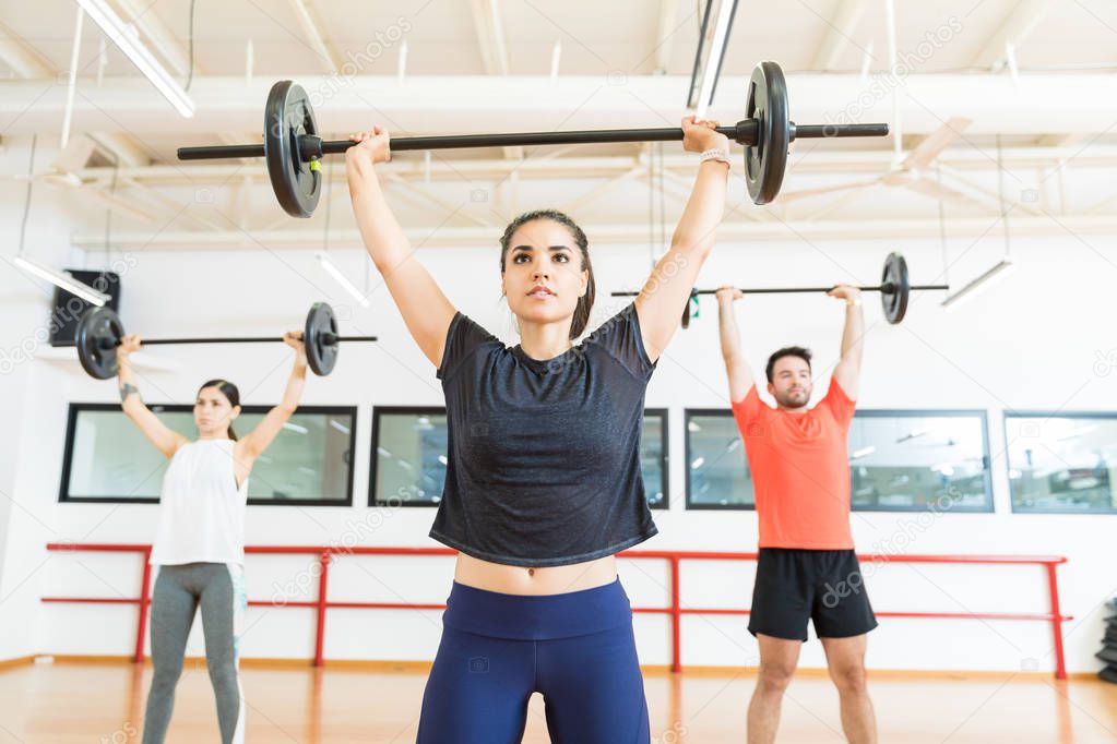 Strong young female lifting barbell while standing in health club