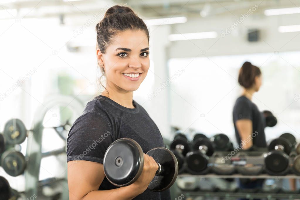 Portrait of female client smiling while doing dumbbell exercise in gym