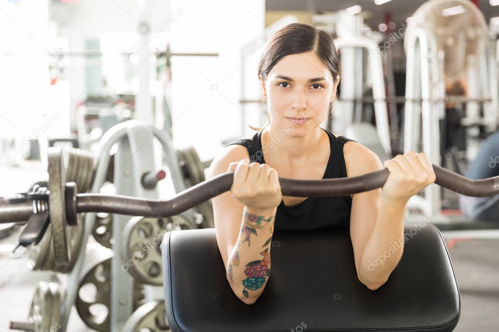 Portrait of confident female athlete lifting barbell curl on exercise machine in gym