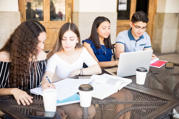 Group of four teenage people sitting at outdoors restaurant making notes and using lapto