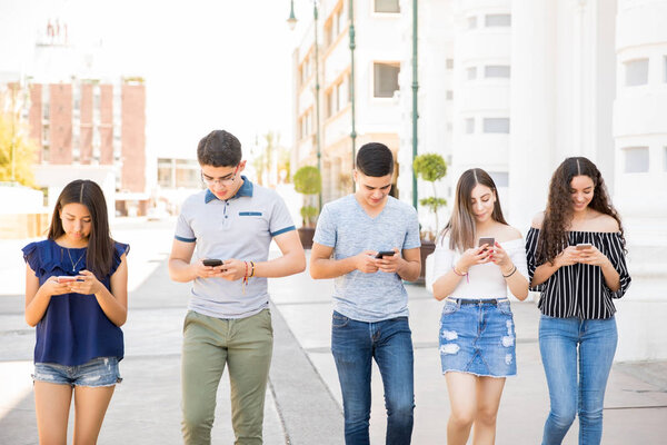 Group of five young boys ands girls walking outdoors in city with their smart phone