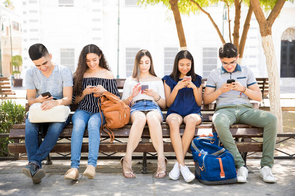 Portrait of teenage boys and girls with backpacks sitting outdoors on bench using their mobile phone