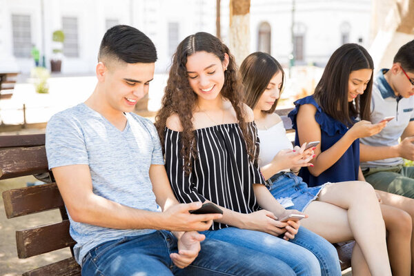 Teenage boy showing something to his friend on smart phone while sitting on bench with other friends social networking