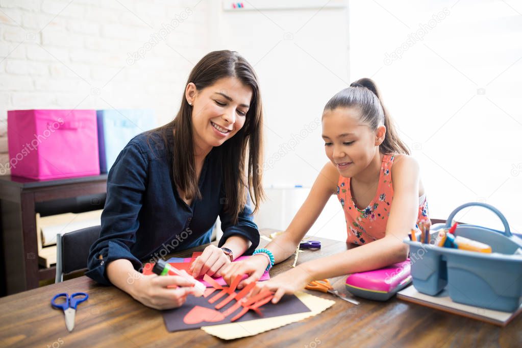 Mid adult mother and daughter gluing paper shape at table in home