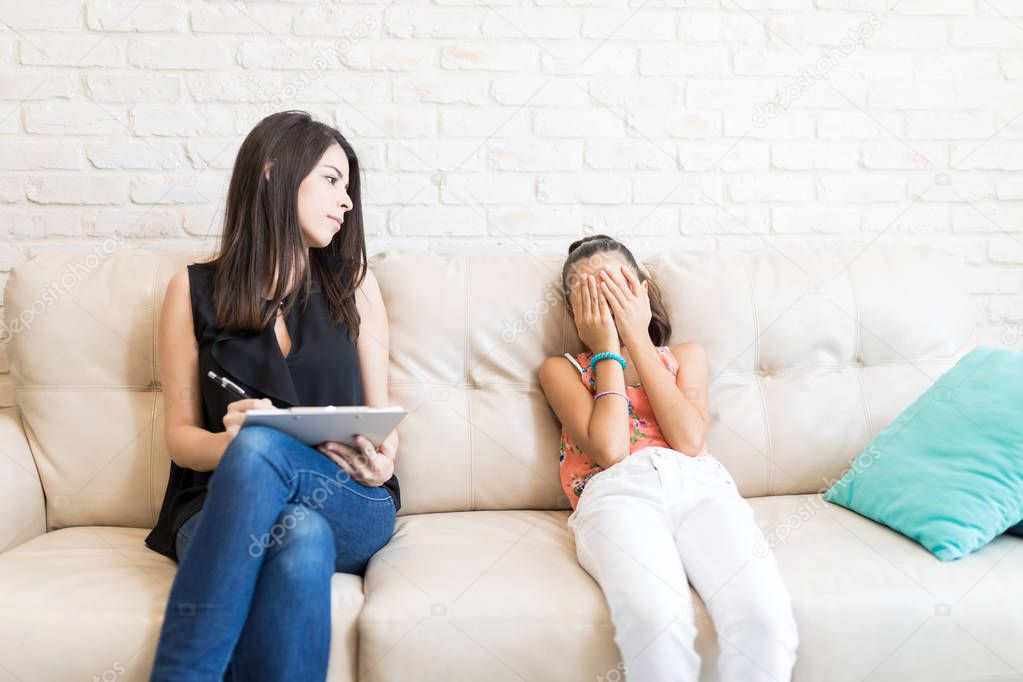 Female psychologist looking at sad girl covering face with hands while sitting on sofa at home
