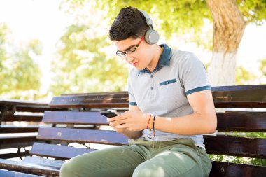 Açık havada ile telefon ve giyen kulaklık müzik dinleme bankta oturan genç latin çocuk portresi