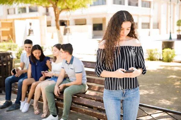 漂亮的年轻女孩的肖像站在户外使用她的智能手机与一群十几岁的青少年坐在后面的长凳上 — 图库照片