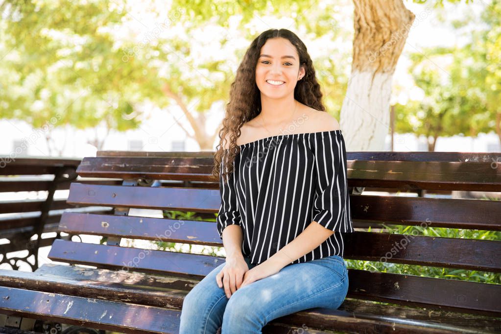 Portrait of beautiful teenage girl sitting on bench outdoors and smiling