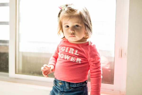 Backlit portrait of a beautiful baby girl standing close to a window at home and making eye contact