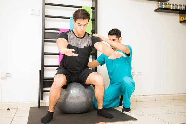 Male physiotherapist helping man balancing on exercise ball in hospital