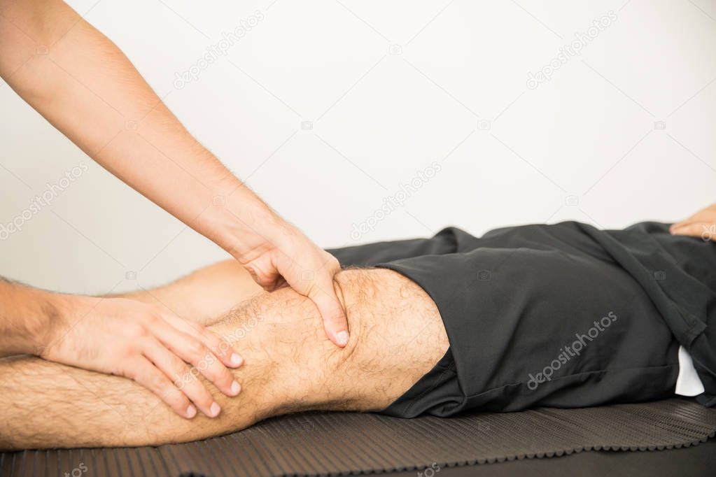 Hands of physiotherapist applying pressure to massage young man's knee in hospital