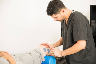 Young male physical therapist placing electrodes on man's knee in hospital clipart