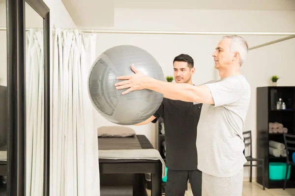 Focused physiotherapist assisting elderly man lifting exercise ball in hospital