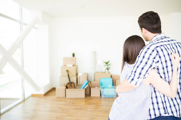 Rear view of mid adult owners embracing each other while looking at moving boxes in new home