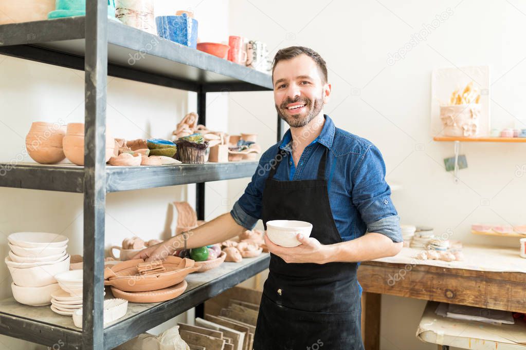 Confident male artisan holding clay bowl while standing by shelves in pottery workshop