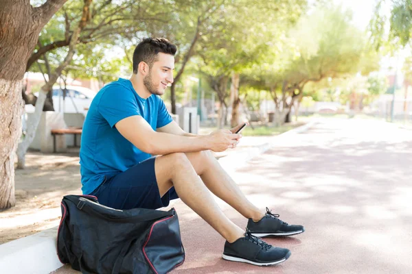 在公园里坐在运动袋上的男性赛跑者的全长使用智能手机 — 图库照片