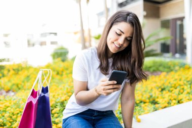Çanta alışveriş merkezi dışında tarafından otururken cep telefonunda orta yetişkin kadın manifatura gülümseyen