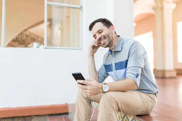 Happy mid adult man using app for listening music on smartphone while sitting at entrance
