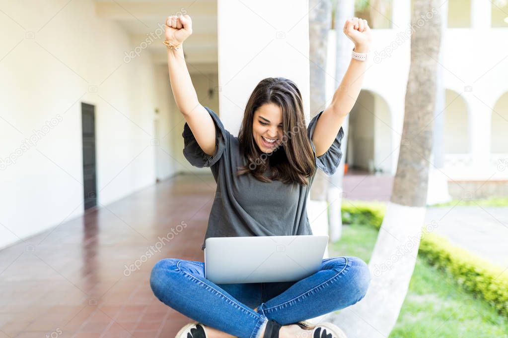 Female with arms raised screaming while using laptop on railing at campus