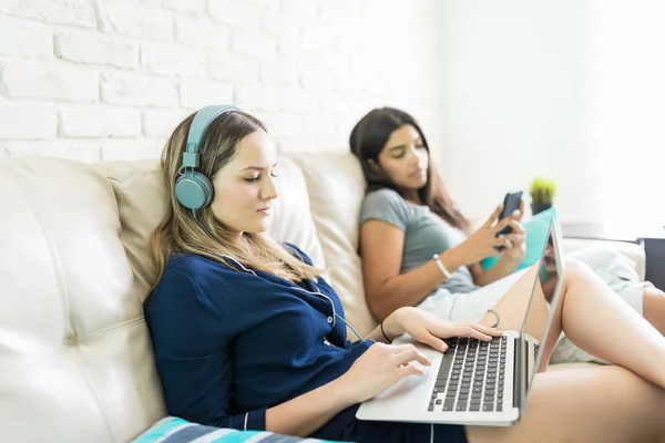 年轻女子在耳机上享受音乐使用笔记本电脑 而朋友在家沙发上休息 — 图库照片
