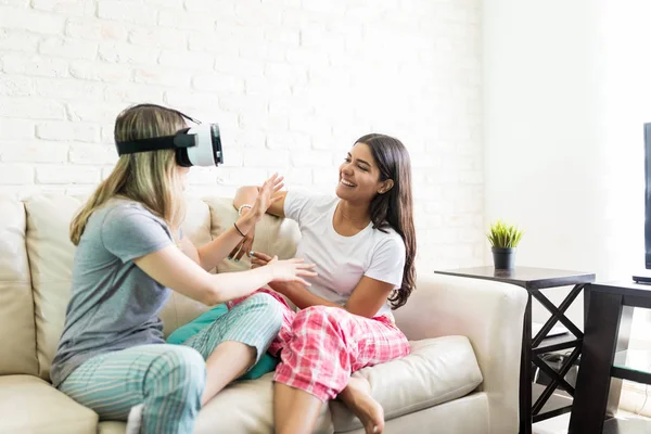 Happy young woman looking at friend wearing virtual reality headset at home