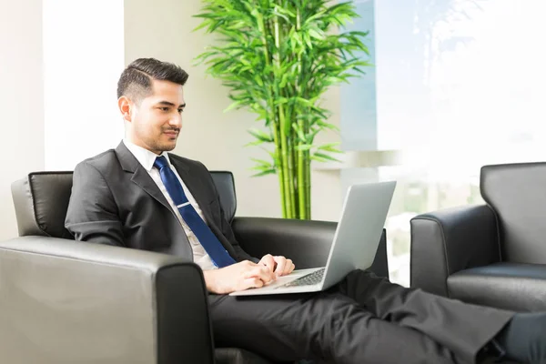 Young male business analyst surfing internet on laptop computer while sitting on chair in lobby of office