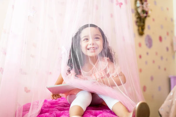 デジタル タブレットの寝室のカーテンを通して見る児の幸せな女性 — ストック写真