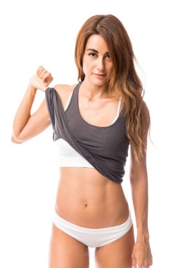 Portrait of female fitness coach picking up tanktop to flaunt flat tummy against white background clipart