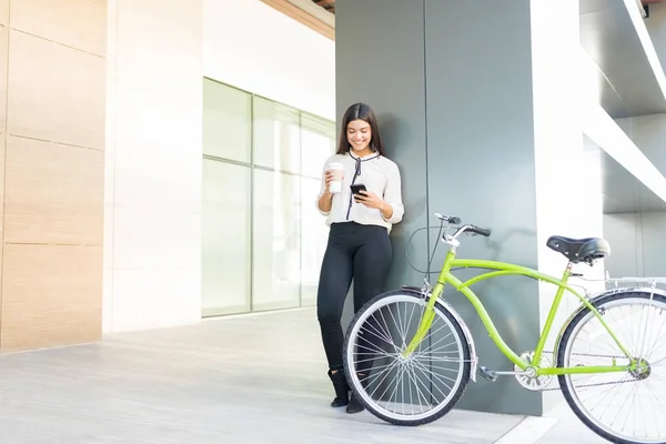 Smiling beautiful commuter holding disposable cup and smartphone by bicycle in office corridor