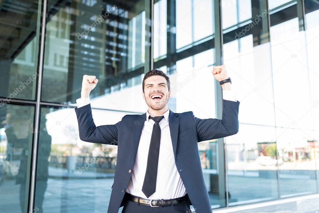 Cheerful Hispanic businessman raising arms and celebrating victory outside office building