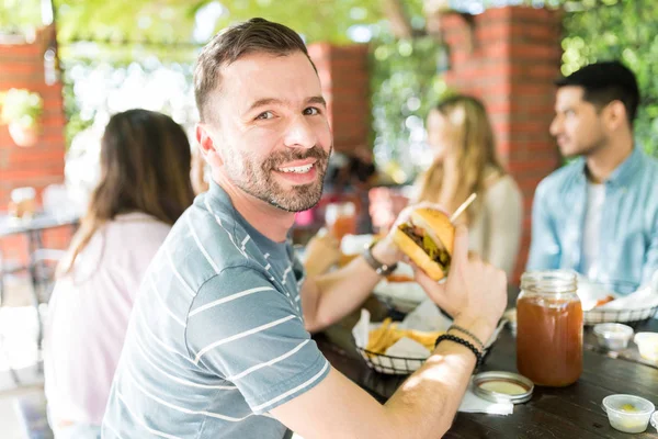 Happy man holding delicious hamburger while sitting with friends