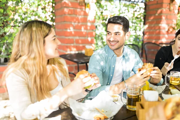 Attractive young man looking at female friend while having cheeseburger at junk food corner