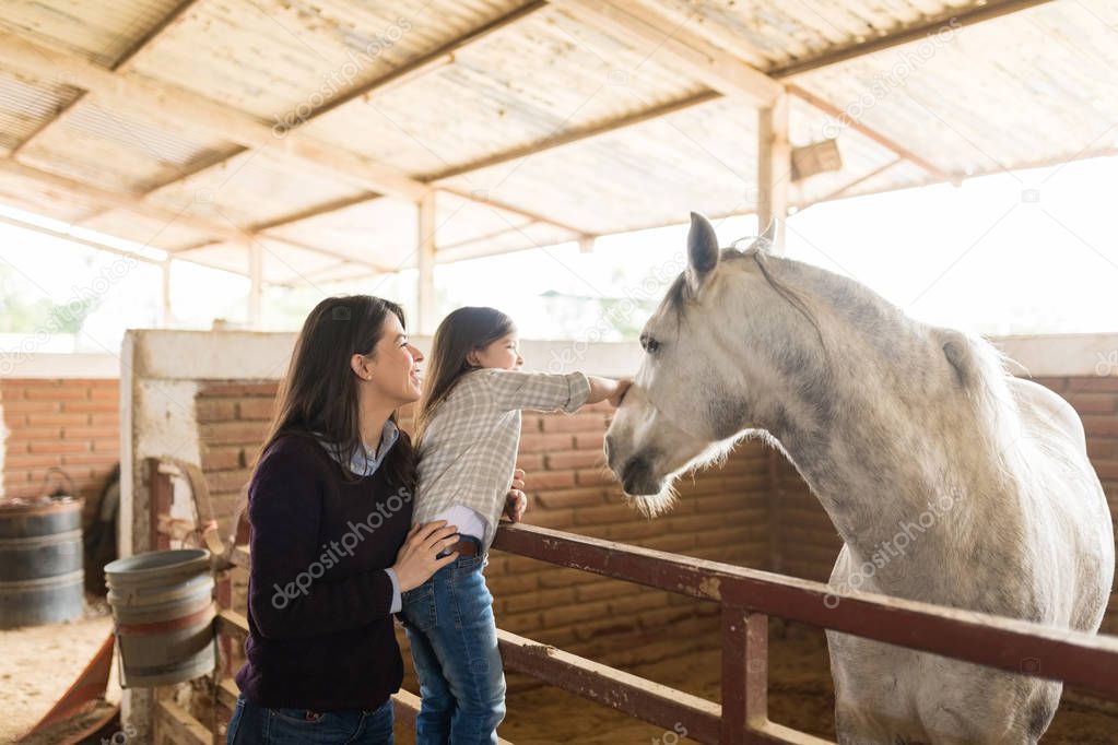 Happy Caucasian mother and daughter showing fondness towards horse in stable