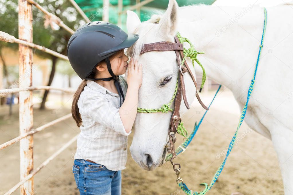 Loving cute girl kissing white horse on head while standing at ranch