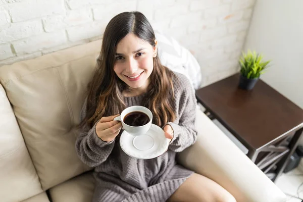 Smiling Hispanic woman in sweater holding coffee cup on sofa