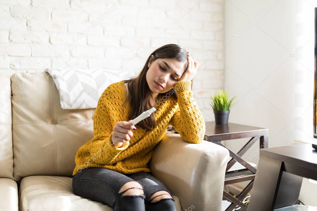 Sad young woman holding pregnancy test kit on sofa at home