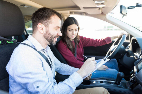 Smiling male examiner writing on clipboard while sitting by new car driver