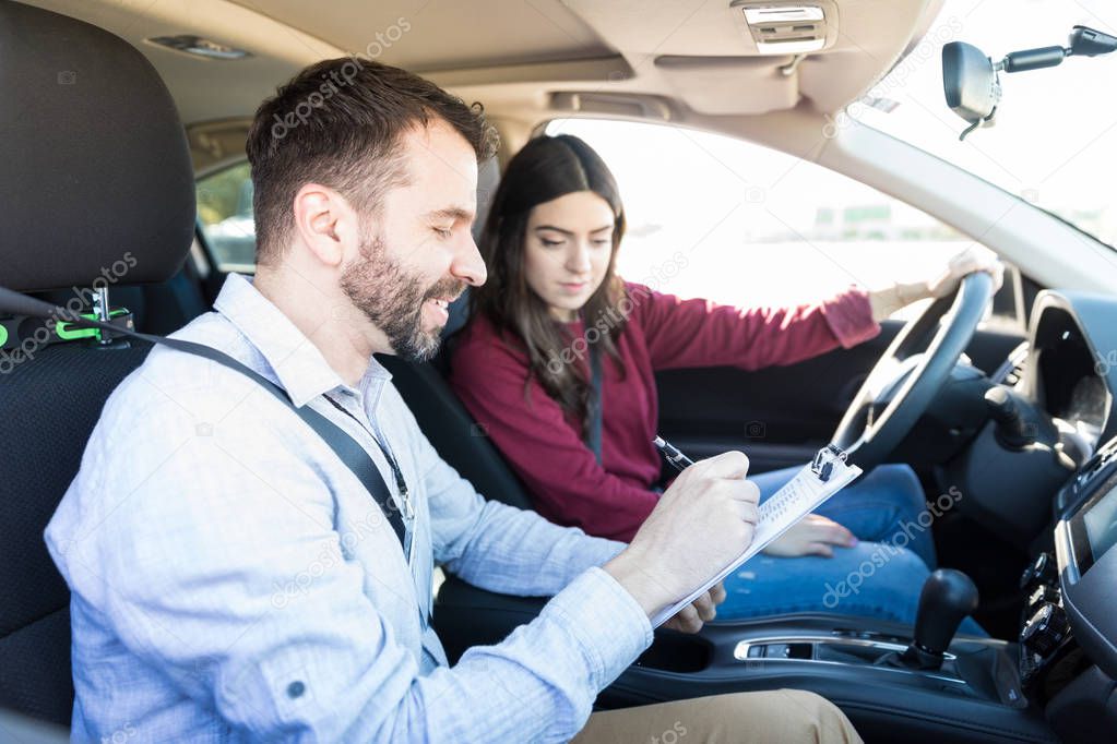 Smiling male examiner writing on clipboard while sitting by new car driver