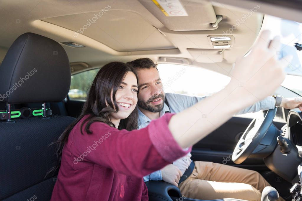 Multiethnic couple smiling while taking selfie with smartphone in new car