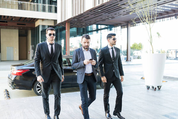 Businessman using smartphone while walking with bodyguards at office campus