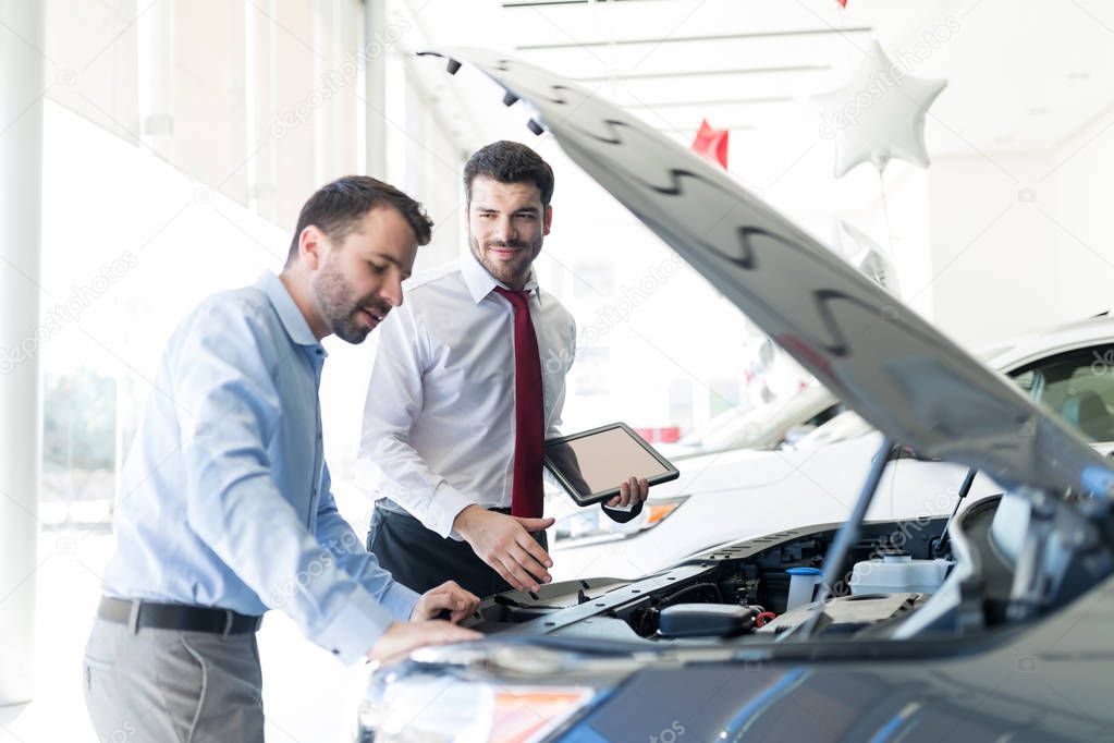 Smiling dealer holding digital tablet while looking at client analyzing car engine