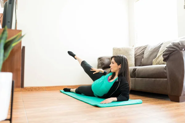 西班牙裔孕妇抬起腿 而斜靠在瑜伽垫在家里 — 图库照片