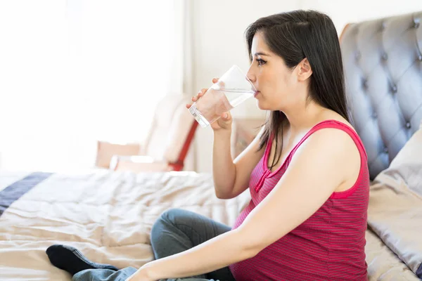 半ば大人の妊娠中の母親は自宅のベッドの上に座っている間はガラスから水を飲む — ストック写真