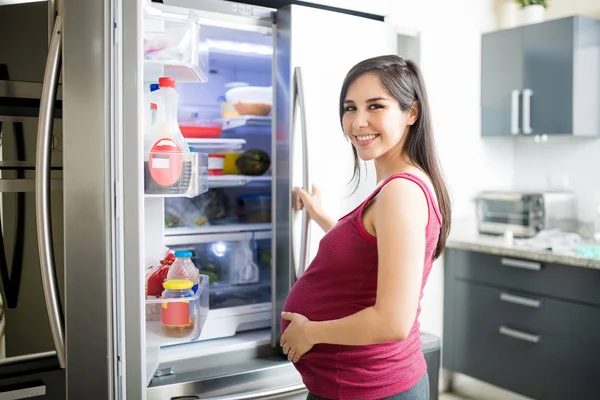 Portrait of beautiful pregnant woman smiling while opening refrigerator in kitchen