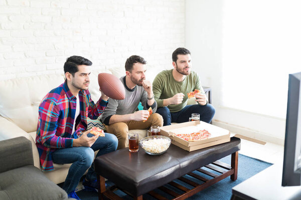 Serious handsome men watching American football match on TV while having some pizza and beer at home