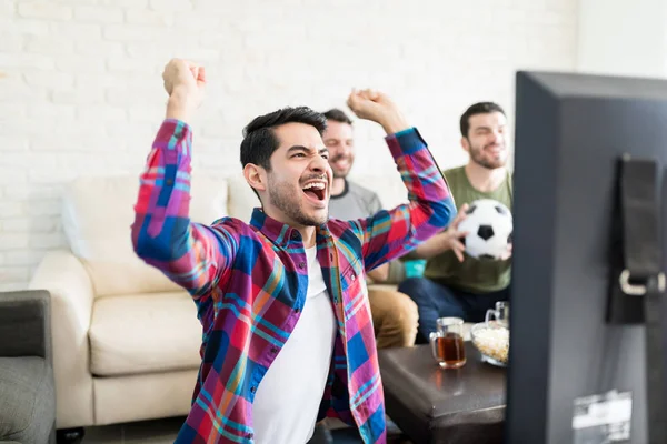 Attractive soccer fan raising arms and screaming in front of TV with friends in background at home