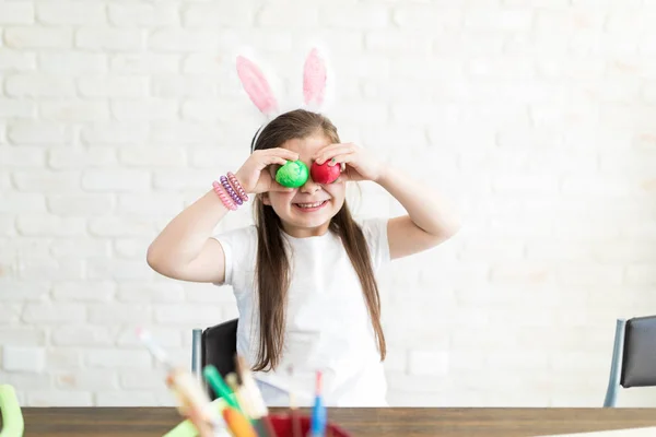 Playful girl covering eyes with Easter eggs while sitting at home