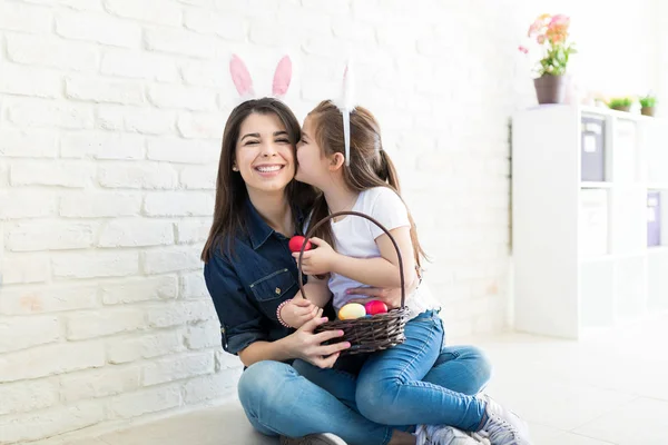 Cute girl with Easter eggs kissing mother wearing bunny ears at home