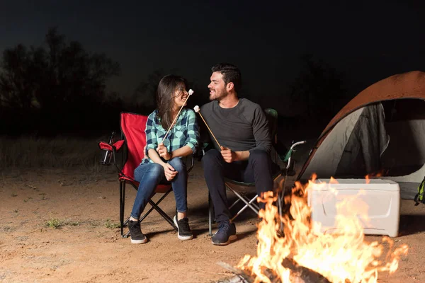 Beautiful couple in love enjoying marshmallows near tent and bonfire in forest